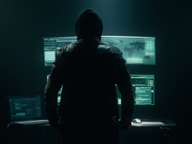 Back view of unrecognizable man in hoodie standing near desk and reading stolen data from computer, monitors in dark room before massive cyber attack on servers.